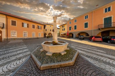 Peveragno, Cuneo, Italy - January 09; 2023: marble fountain with drinking trough on cobblestones in Piazza Santa Maria with ancient palaces decorati with arcade under cloudy sky at sunset
