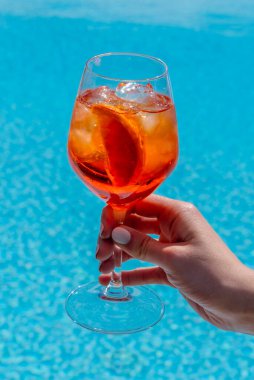 Aperol Spritz alkollü kokteyl. Mavi su havuzu arka planında beyaz tırnak cilası olan bir kadının elinde cam kadehte. 