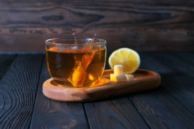 Fotoğrafta, bir bardak çay ahşap bir tepsiye konuyor, yanında da bir dilim limon ve birkaç küp şeker bulunuyor. Arka planda koyu tahta levhalar var.
