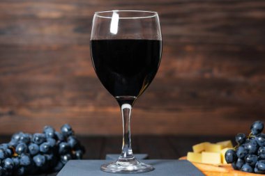 Arka planda üzüm ve peynir demetleri olan ahşap bir masada bir bardak kırmızı şarap. Sıcak ışıklandırma ve kırsal ahşap zemin rahat ve davetkar bir atmosfer yaratır.