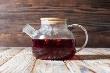 Kırmızı renkli çayla dolu bir kapağı olan cam çaydanlık ahşap bir masada oturur. Arka plandaki ahşap doku rahat bir atmosfer yaratıyor.