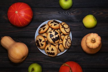  Balkabağı ve elmalarla çevrili, çikolata kabağı yüzlü bir tabak kurabiye. Koyu renk ahşap masa, Cadılar Bayramı için mükemmel bir sonbahar atmosferi ekler..