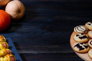 Ahşap bir tabakta ürkütücü tasarımları olan Cadılar Bayramı kurabiyeleri. Etrafı karanlık ahşap bir masada balkabaklarıyla çevrili. Mevsimlik kutlamalar, yemek stili ve sonbahar temaları için ideal.