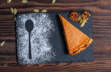 Kaşık, ceviz ve kabak çekirdeği şeklinde pudra şekerli siyah bir tabakta canlı bir portakallı balkabağı dilimi. Sonbahar temalı tasarımlar, yemek blogları ve menüler için ideal. 