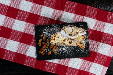 A festive Christmas stollen cake decorated with powdered sugar and dried fruits, sitting on a black slate plate against a red and white checkered tablecloth. clipart