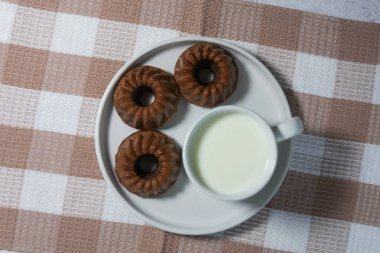  A top-down view of three mini chocolate bundt cakes and a cup of milk on a white plate, placed on a checkered tablecloth. A simple and cozy food composition. clipart