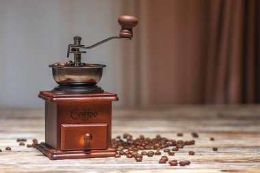 Metal saplı eski bir kahve öğütücüsü ve kahve çekirdeği dolu bir kase tahta bir masanın üzerinde duruyor. Kahve çekirdekleri yakınlarda..