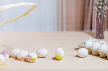 Tahta bir masanın üzerinde, karton bir paketin yanında çizimleri olan parlak renkli Paskalya yumurtaları. Arka planda yeşil dallı bir sepet ve vazo var. Bahar tatilinin atmosferi.