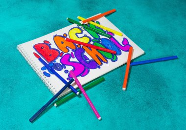 Canlı Okula Dönüş dizaynı, turkuaz bir arka planda renkli işaretlerle çevrili spiral bir deftere çizilmiş. Eğitim, kırtasiye veya sınıf temaları için neşeli kompozisyon.