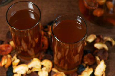 Kurutulmuş meyve kompostosu ile dolu iki bardak kayısılar, elmalar ve kahverengi dokulu kuru üzümlerle çevrili. Doğal malzemelerle ev yapımı sıcak bir içecek.