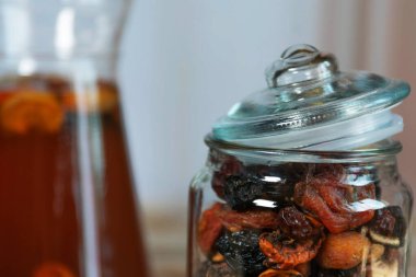 Kayısı ve kuru üzüm gibi çeşitli kurumuş meyvelerle dolu eğimli kapaklı cam bir kavanoz. Arka plandaki kompote sürahisi tablo ayarına karşıtlık ekler.