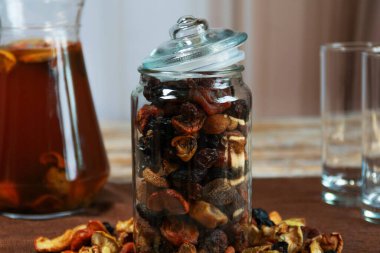 Kurutulmuş meyvelerle dolu cam kavanoz daha fazla meyve ve bardakla çevrili bir masada açık duruyor. Sağlıklı atıştırmalıklar, ev yapımı tarifler ve sonbahar temaları için mükemmel..