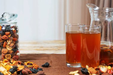 Bir kavanoz elma, kayısı ve kuru eriğin yanında kurutulmuş meyve kompostosu dolu sürahi ve bardaklar. Kahverengi kumaşla ahşap yüzeye kurulmuş rahat bir yer. Doğal ev yapımı içecek sahnesi.