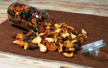 Cam kavanoz devrilmiş, ahşap bir masanın üzerine bir sürü kurutulmuş meyve dökülmüş. Karışımda elmalar, kayısılar ve kuru üzümler kırsal ve organik bir görüntü oluşturur..