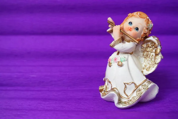 Guardian angel plays the flute. Figurine of a red-haired girl in a white dress with wings and a pipe. New Year or Christmas holiday background. copyspace. Purple background. Free space for text