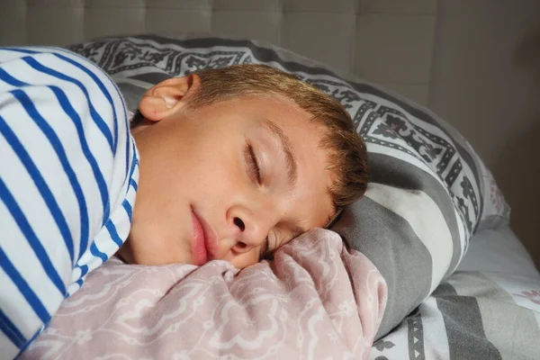 A beautiful caucasian boy of 10 years old with blond hair, dressed in striped pajamas, sleeps on a bed with a fluffy blanket, hugging a pillow. Soft light of the morning sun streams through the window