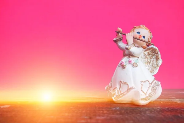 Guardian angel plays the flute. Figurine of a red-haired girl in a white dress with wings and a pipe. New Year or Christmas holiday background. Copy space. Pink wooden background. Sunshine or ray