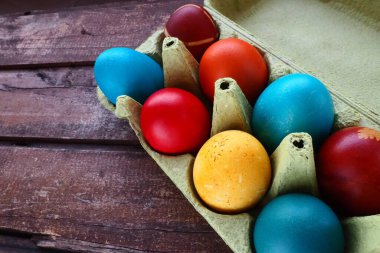 Multi-colored eggs painted with gouache and onion peel in a cardboard box on a wooden table background. Ten boiled eggs. Postcard poster for Easter. Easter holiday. Red, blue, yellow, brown eggs.