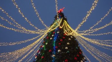 Shiny balls on the New Years city tree. Glowing garlands hang over the Christmas tree. Outdoor winter decoration. Festive design of the city square. Garlands blink and sway in the wind. A red star.