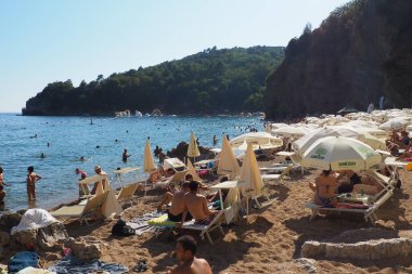 Budva, Montenegro, Mogren beach. August 15 2022 Sand and pebble beach near high cliffs. Beach umbrellas, cafes, deck chairs. People relax, swim, enjoy and sunbathe. Tourism business in the Balkans