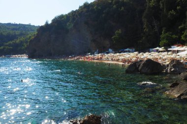 Budva, Karadağ, Mogren plajı. 15 Ağustos 2022. Yüksek kayalıkların yakınındaki kum ve çakıl taşı plajı. Plaj şemsiyeleri, kafeler, şezlonglar, şezlong. İnsanlar rahatlar, yüzer, güneşlenir. Balkanlar 'da turizm sektörü