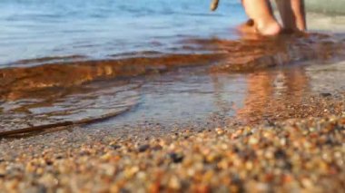Waves of clear water crash against the sandy beach. The child stands ankle-deep in water, plays with a stick. Babys legs and the edge of a blue shirt. Golden hour, sunset. Sea, ocean, lake or river