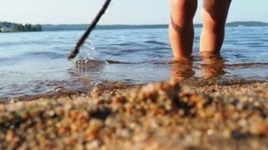 Waves of clear water crash against the sandy beach. The child stands ankle-deep in water, plays with a stick. Babys legs and the edge of a blue shirt. Golden hour, sunset. Sea, ocean, lake or river