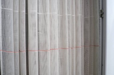 White coarse cotton or linen curtains made of weaves of thin threads. Simple compact economical apartment interior. Window decoration in the room. Red stripe