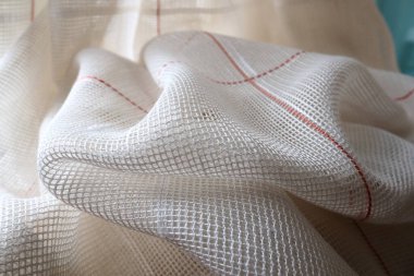 White coarse cotton or linen curtains made of weaves of thin threads. Simple compact economical apartment interior. Window decoration in the room. Red stripe. The fabric is folded in soft waves.