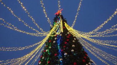 Shiny balls on the New Years city tree. Glowing garlands hang over the Christmas tree. Outdoor winter decoration. Festive design of the city square. Garlands blink and sway in the wind. A red star.