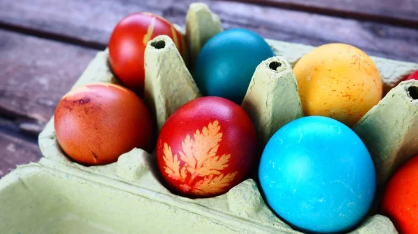 Multi-colored eggs painted with gouache and onion peel in a cardboard box on a wooden table background. Ten boiled eggs. Postcard poster for Easter. Easter holiday. Red, blue, yellow, brown eggs.
