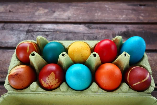 Multi-colored eggs painted with gouache and onion peel in a cardboard box on a wooden table background. Ten boiled eggs. Postcard poster for Easter. Easter holiday. Red, blue, yellow, brown eggs.