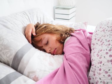 8 yaşında, sarı saçlı, pembe pijamalı, tüylü bir battaniyeyle yatakta uyuyan, yastığına sarılan güzel bir beyaz kız. Sabah güneşinin yumuşak ışığı pencereden içeri akıyor.