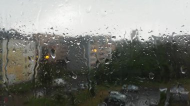 Yağmurlu bir günde cam pencereye yağmur damlaları düşer. Yukarıdan su damlıyor. Arka plan bulanık. Şehirde kötü hava var. Binalar, evler, pencerenin dışındaki arabalar. Meteoroloji ve hava tahmini. Bulutluluk.