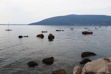 Herceg Novi, Karadağ. Deniz tatili. Adriyatik Denizi 'nde tekneler yüzer. Akdeniz seyahat işi. Yelkenliler ve dağlar. Tuzlu suda küçük dalgalanmalar. Dalgalarda gökyüzü yansıması. Siyah taşlar.