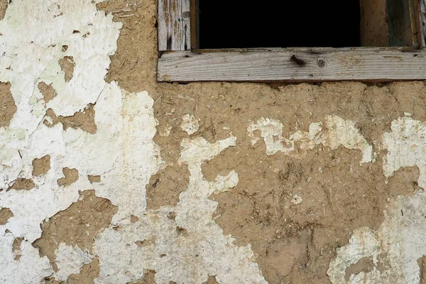 The wall of a residential old house built with bricks made from mud, cow dung and straw. Peeling plaster. Serbia, ancient building technology. Texture of plaster and bricks. Window with wooden frame