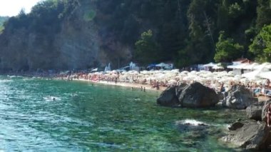 Budva, Karadağ, Mogren plajı. 15 Ağustos 2022. Yüksek kayalıkların yakınındaki kum ve çakıl taşı plajı. Plaj şemsiyeleri, kafeler, şezlonglar, şezlong. İnsanlar rahatlar, yüzer, güneşlenir. Balkanlar 'da turizm sektörü