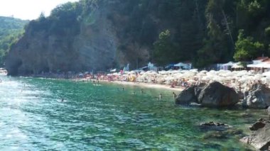 Budva, Karadağ, Mogren plajı. 15 Ağustos 2022. Yüksek kayalıkların yakınındaki kum ve çakıl taşı plajı. Plaj şemsiyeleri, kafeler, şezlonglar, şezlong. İnsanlar rahatlar, yüzer, güneşlenir. Balkanlar 'da turizm sektörü