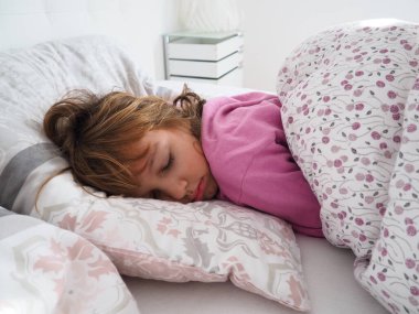 8 yaşında, sarı saçlı, pembe pijamalı, tüylü bir battaniyeyle yatakta uyuyan, yastığına sarılan güzel bir beyaz kız. Sabah güneşinin yumuşak ışığı pencereden içeri akıyor.