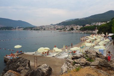 Herceg Novi, Karadağ, 09 Ağustos 2022 beton plakalı popüler çakıl taşı plajı. Güneş pansiyonları, oturma yerleri ve şemsiyeler. Turistler güneşlenir. Adriyatik denizi ve dağları. Deniz kenarındaki kafe ve sahil servisi.