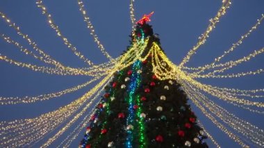 Shiny balls on the New Years city tree. Glowing garlands hang over the Christmas tree. Outdoor winter decoration. Festive design of the city square. Garlands blink and sway in the wind. A red star.