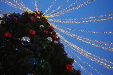 Multi-colored glass shiny balls on the New Years city tree. Glowing garlands hang over the Christmas tree. Outdoor winter decoration. Festive design of the city square. Red, blue and yellow ball.