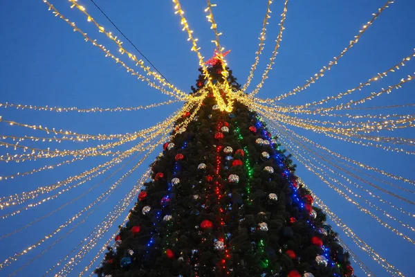 Multi-colored glass shiny balls on the New Years city tree. Glowing garlands hang over the Christmas tree. Outdoor winter decoration. Festive design of the city square. Red, blue and yellow ball.