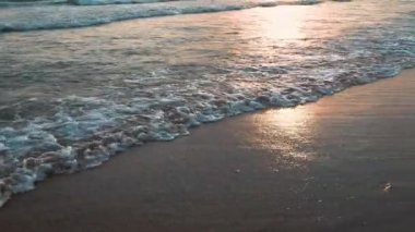 Sakin dalgalar kumlu sahilde yuvarlanıyor. Denizde ya da okyanusta gün batımı. Su hareketi. Dalgalar köpükten köpürüyor. Turist cenneti. Seyahat Sahili Teması. Güneşin dalgalardaki yansıması