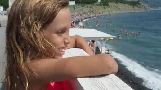 一个9岁的白种人女孩 长发望着海滩 全身都是日光浴和游泳的人 然后转身对着摄像机 用张开的嘴和皱巴巴的鼻子做出可怕的鬼脸 — 图库视频影像