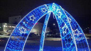Petrozavodsk Karelia Rusya 01.12.24 Yeni Yıl süsleri ve yanıp sönen ışıklarla parıldayan dekoratif kemer. Mavi ve beyaz çelenkler. Tatil dekoru. Retro çan. Kış akşamı, alışveriş merkezi, arabalar..