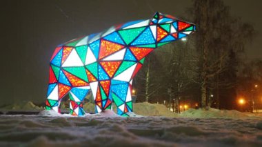 Ayı çelengi. Çok renkli, ışıl ışıl parlayan yeni yıl ayısı geceleri kar üzerinde. Kurulum figürü, tatilde sokakları dekore etmek için mimari form. Kırmızı, mavi, yeşil, beyaz üçgen..
