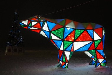 Ayı çelengi. Çok renkli, ışıl ışıl parlayan yeni yıl ayısı geceleri kar üzerinde. Kurulum figürü, tatilde sokakları dekore etmek için mimari form. Kırmızı, mavi, yeşil, beyaz üçgen..