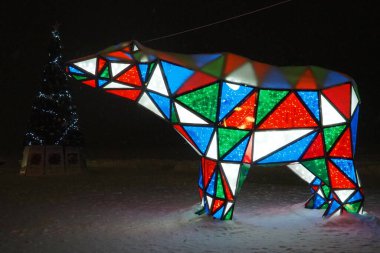 Ayı çelengi. Çok renkli, ışıl ışıl parlayan yeni yıl ayısı geceleri kar üzerinde. Kurulum figürü, tatilde sokakları dekore etmek için mimari form. Kırmızı, mavi, yeşil, beyaz üçgen..