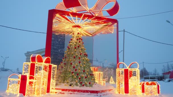 金色の赤いガーランドの電球から成っている明るいクリスマス ツリーおよび大きい弓が付いているギフト用の箱の形の建築様式 都市の通りや広場の装飾を祝う クリスマスと新年 — ストック動画